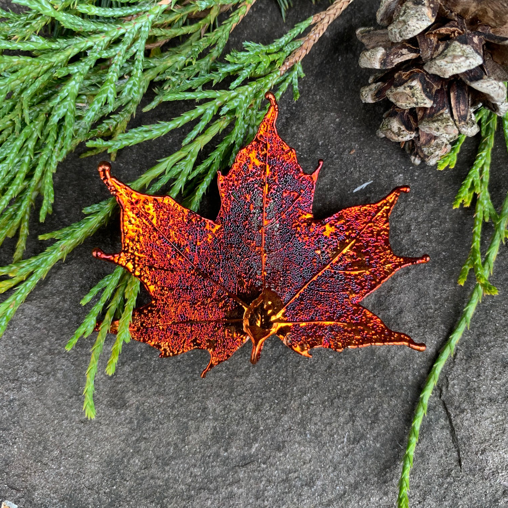 A small metal coated maple leaf sits on a stone background. The metal has an iridescent finish and sparkles in several shades of orange and red. At the top of the picture are decorative evergreen leaves and a pine cone.