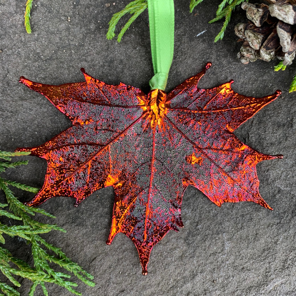 A large metal coated maple leaf sits on a stone background. The metal has an iridescent finish, and sparkles in several shades of orange and red. A green ribbon is attached to the stem end of the leaf.  Around the picture are decorative evergreen leaves and a pine cone.