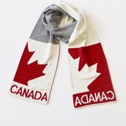 A red, white, and grey Canada scarf. The middle of the scarf is twisted up, but the ends light flat to show off the designs on the scarf. The design is of half a maple leaf and the word Canada. This reversible design is red on white on one side of the scarf, and white on red on the other.