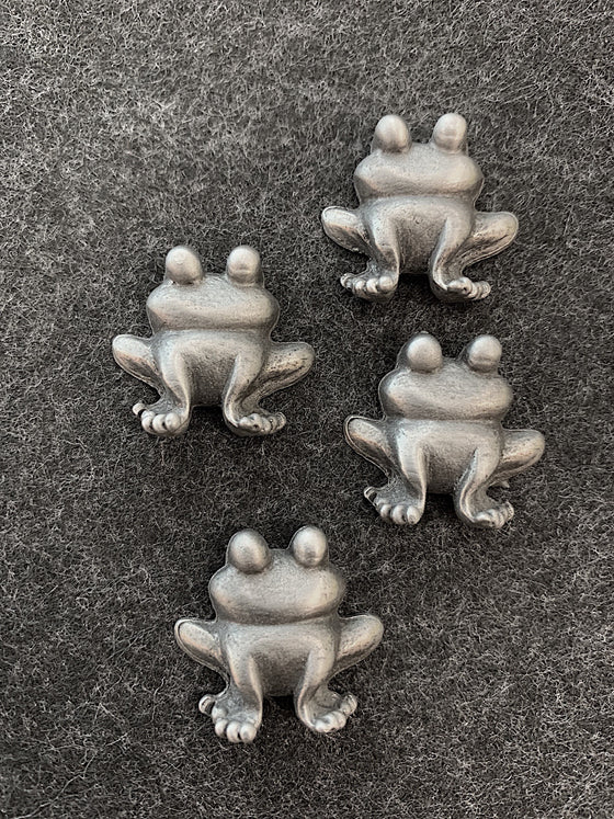four pewter magnets in the shape of frogs facing forward with large cartoonish eyes on the tops of their heads. 
