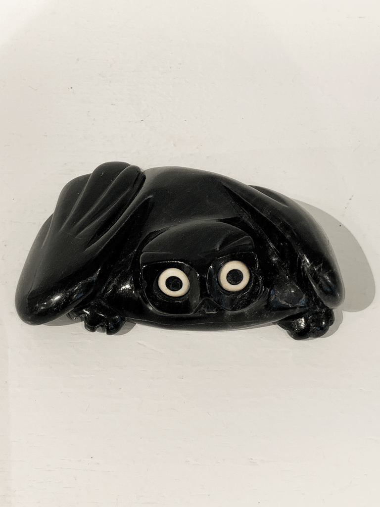 A soapstone carving of a "pancake owl" ( title due to the sculpture's flat quality) by Joanasie Manning. This piece is made from dark black stone with some marbled grey patterning throughout. In this photograph, a front view of the owl is shown.