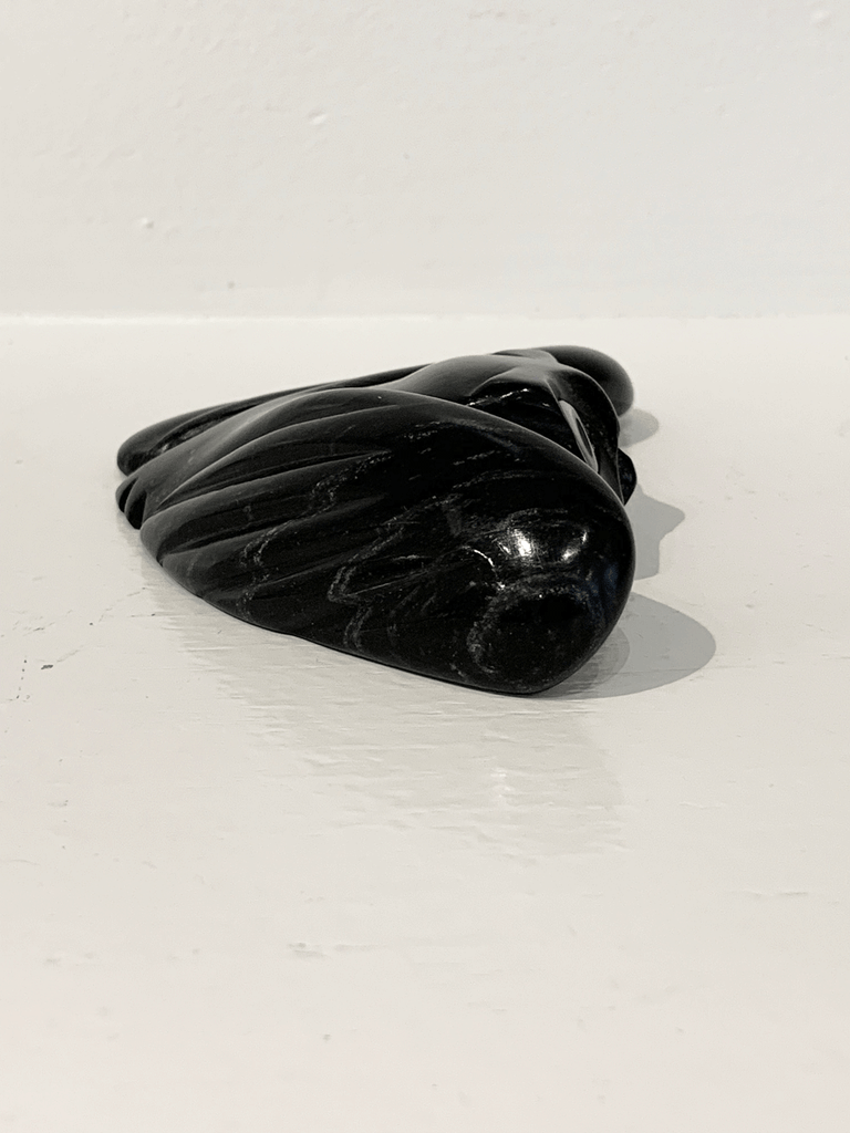 A soapstone carving of a "pancake owl" ( title due to the sculpture's flat quality) by Joanasie Manning. This piece is made from dark black stone with some marbled grey patterning throughout. In this photograph, a side view of the owl is shown.