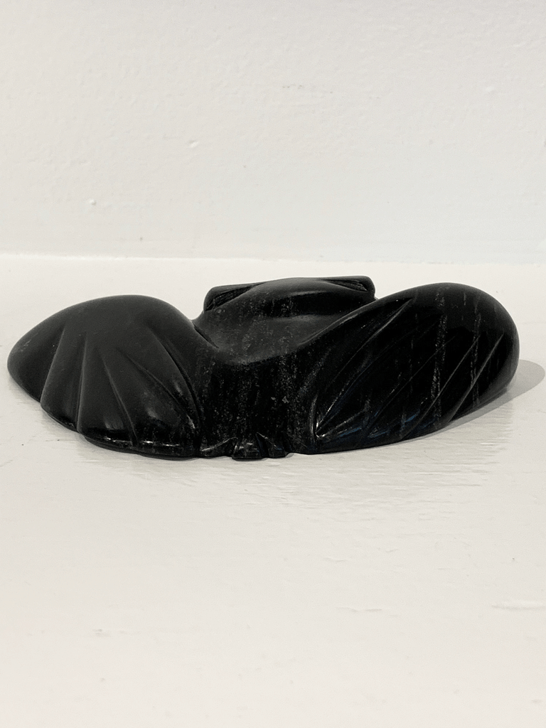 A soapstone carving of a "pancake owl" ( title due to the sculpture's flat quality) by Jordan Kelly. This piece is made from dark black stone with some marbled grey patterning throughout. In this photograph, a back view of the owl is shown.