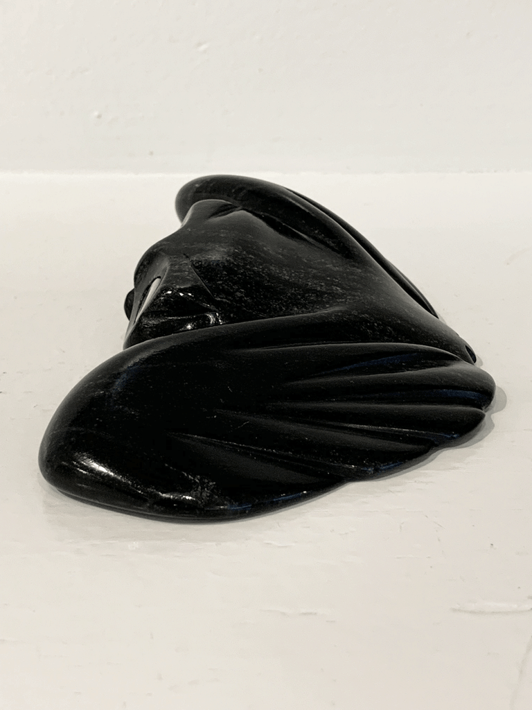 A soapstone carving of a "pancake owl" ( title due to the sculpture's flat quality) by Jordan Kelly. This piece is made from dark black stone with some marbled grey patterning throughout. In this photograph, a side view of the owl is shown.