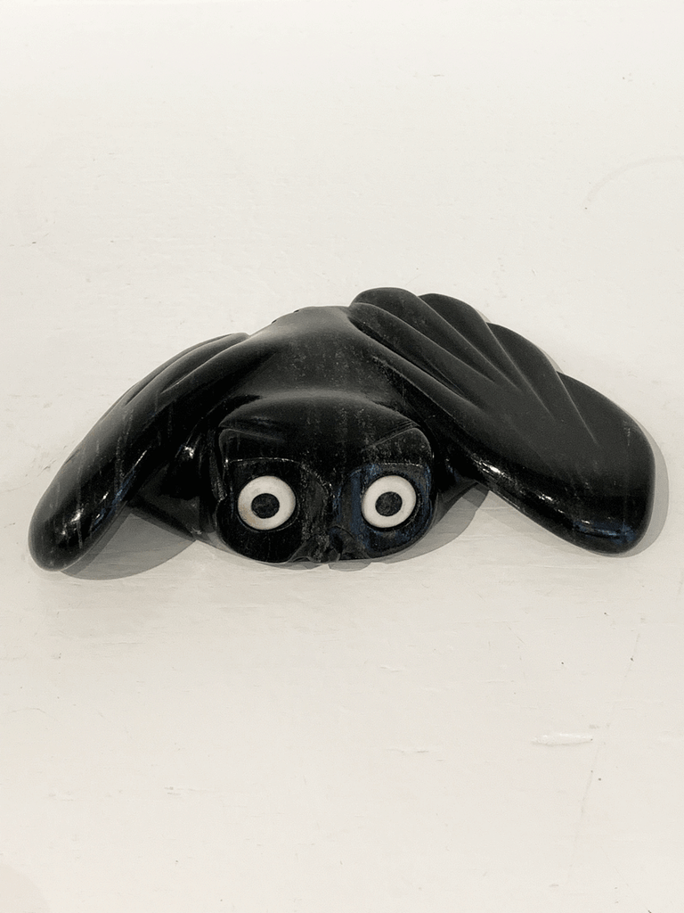 A soapstone carving of a "pancake owl" ( title due to the sculpture's flat quality) by Joanasie Manning. This piece is made from dark black stone with some marbled grey patterning throughout. In this photograph, a front view of the owl is shown.