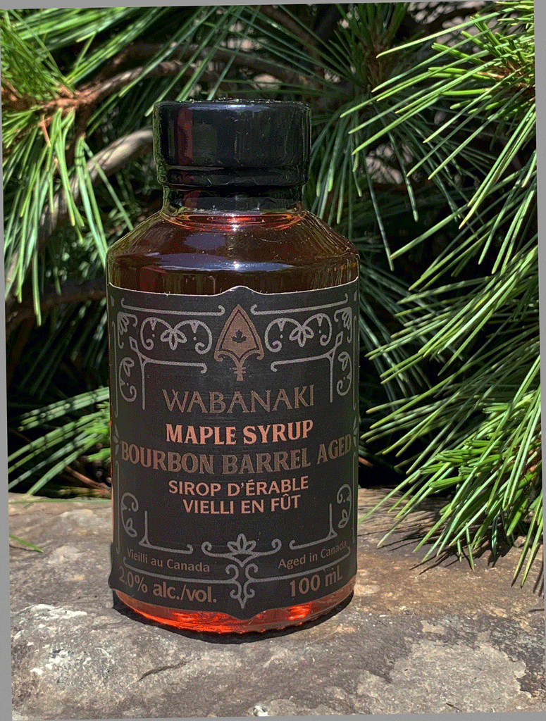 A clear bottle of beautiful amber maple syrup. In gold lettering on a black label, it reads "Wabanaki Maple Syrup Bourbon Barrel Aged" and "Sirop D'erable Vieilli en Fut de Bourbon." In smaller letters at the bottom, it reads "Vieilli au Canada/Aged in Canada," "2.0% alc./vol." and "100 ml."