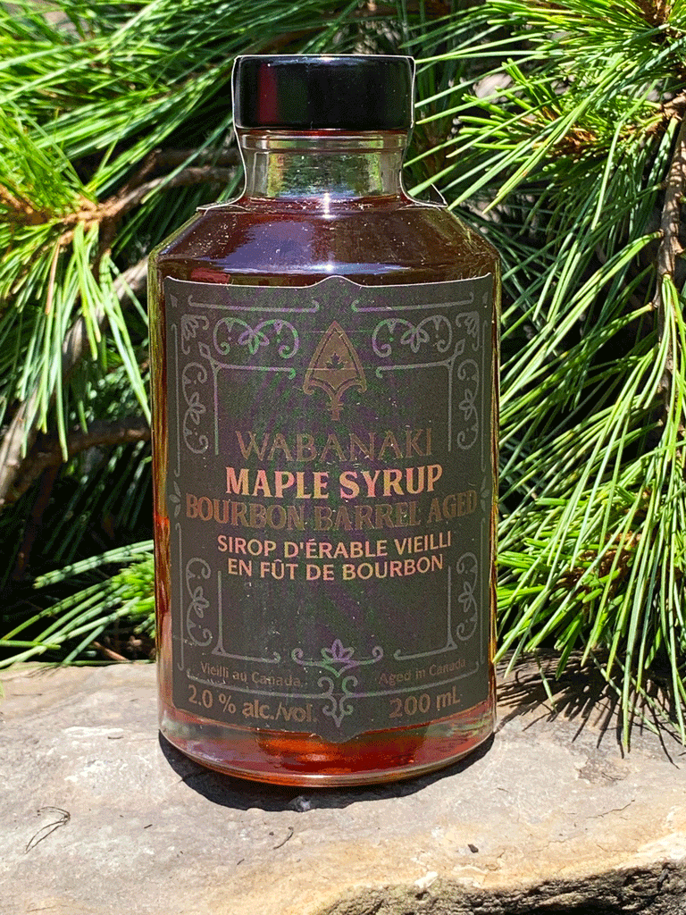 A clear bottle of beautiful amber maple syrup. In gold lettering on a black label, it reads "Wabanaki Maple Syrup Bourbon Barrel Aged" and "Sirop D'erable Vieilli en Fut de Bourbon." In smaller letters at the bottom, it reads "Vieilli au Canada/Aged in Canada," "2.0% alc./vol." and "200 ml."
