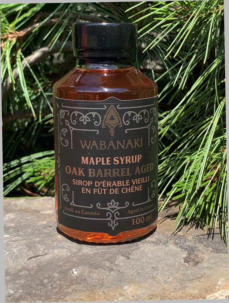 A clear bottle of beautiful amber maple syrup. In gold lettering on a black label, it reads "Wabanaki Maple Syrup Oak Barrel Aged" and "Sirop D'erable Vieilli en Fut de Chene." In smaller letters at the bottom, it reads "Vieilli au Canada/Aged in Canada," "2.0% alc./vol." and "100 ml."