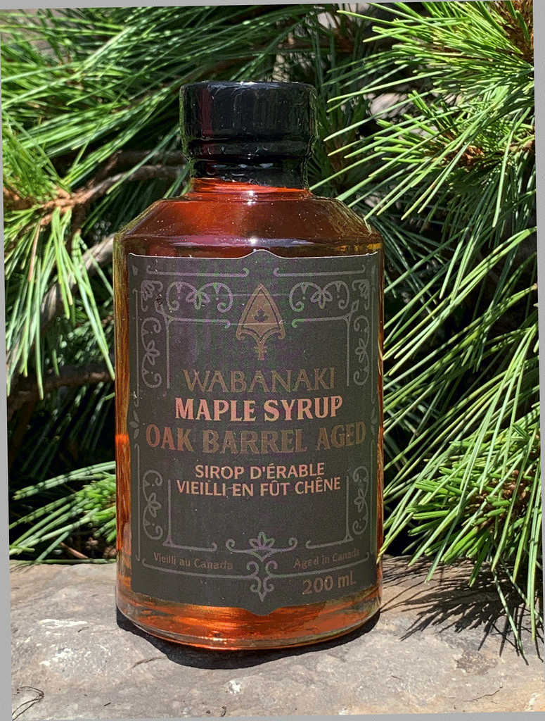 A clear bottle of beautiful amber maple syrup. In gold lettering on a black label, it reads "Wabanaki Maple Syrup Oak Barrel Aged" and "Sirop D'erable Vieilli en Fut de Chene." In smaller letters at the bottom, it reads "Vieilli au Canada/Aged in Canada," "2.0% alc./vol." and "200 ml."