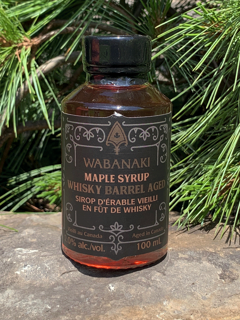 A clear bottle of beautiful amber maple syrup. In gold lettering on a black label, it reads "Wabanaki Maple Syrup Whisky Barrel Aged" and "Sirop D'erable Vieilli en Fut de Whisky." In smaller letters at the bottom, it reads "Vieilli au Canada/Aged in Canada," "2.0% alc./vol." and "100 ml."