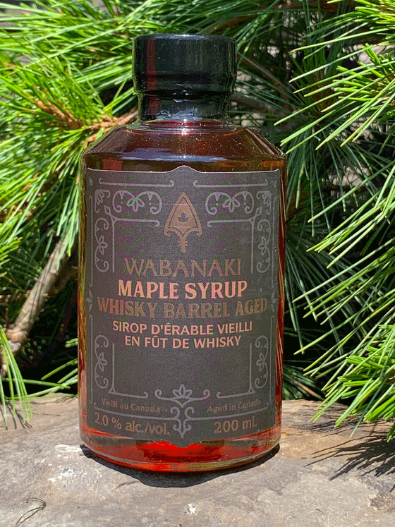 A clear bottle of beautiful amber maple syrup. In gold lettering on a black label, it reads "Wabanaki Maple Syrup Whisky Barrel Aged" and "Sirop D'erable Vieilli en Fut de Whisky." In smaller letters at the bottom, it reads "Vieilli au Canada/Aged in Canada," "2.0% alc./vol." and "200 ml."